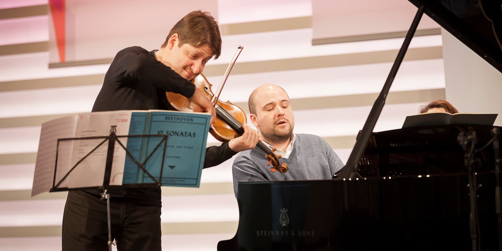 A violinist and a pianist perform together on stage. In front of the violinist, there is a music stand with Beethoven's Sonatas.