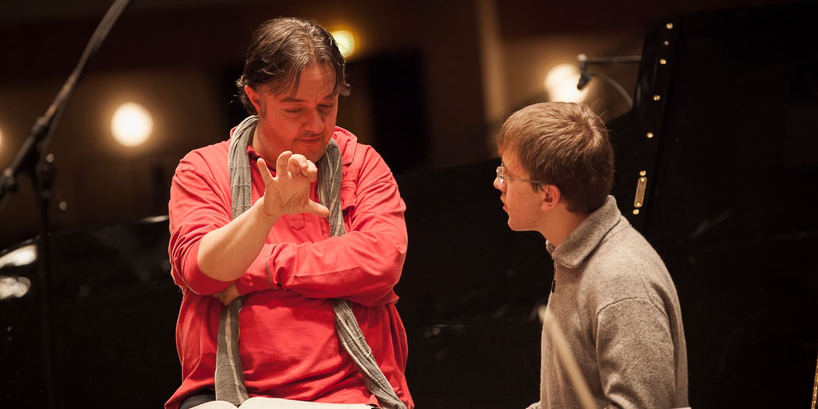 A man in a red shirt and scarf is gesturing while discussing with a younger man in a gray sweater.