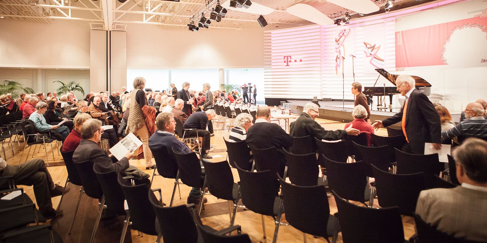 People sitting and standing in a large, well-lit event room in front of a stage with a grand piano.