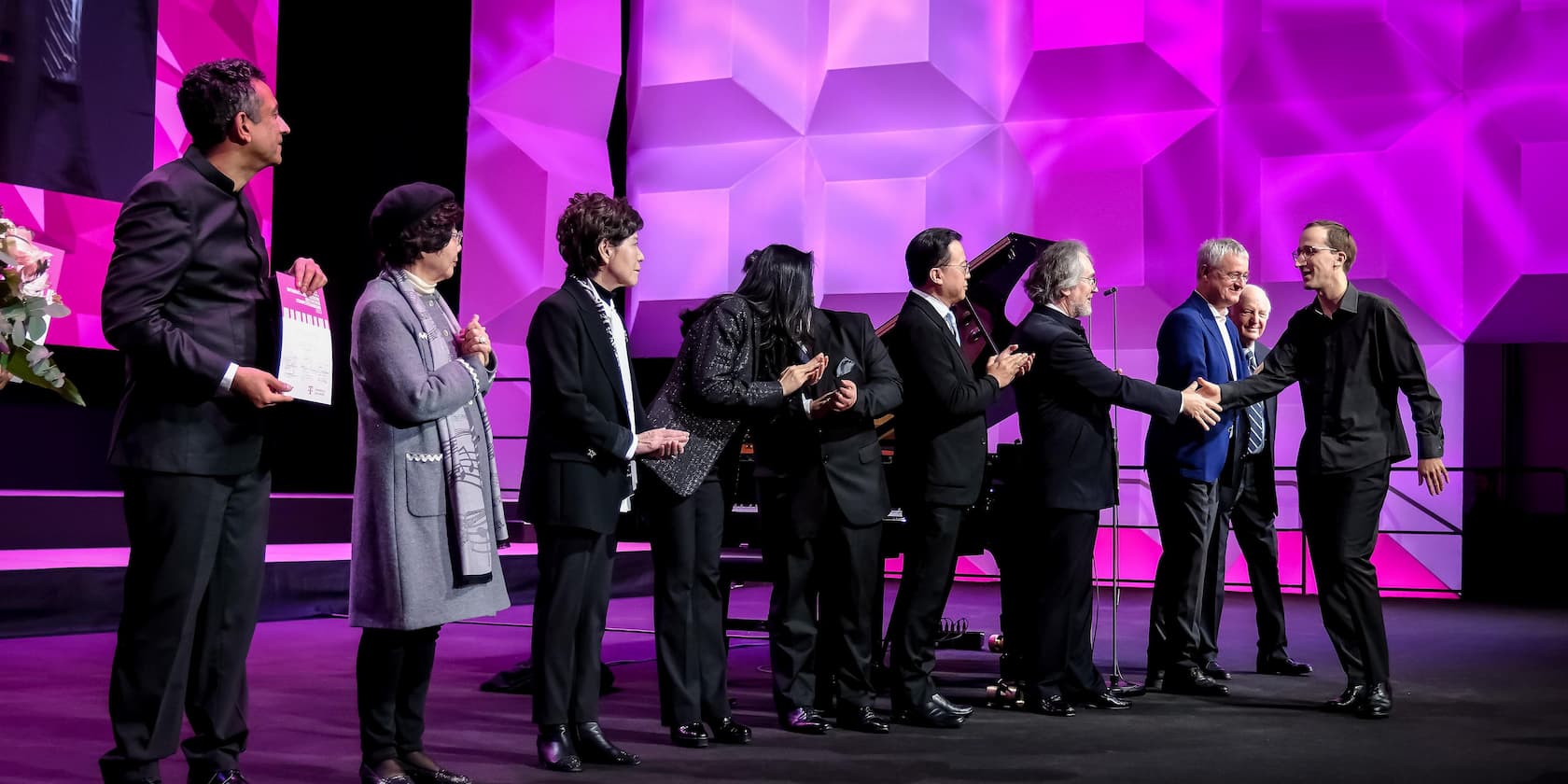 A group of people standing on a stage and applauding while two men shake hands. The background is lit in purple.