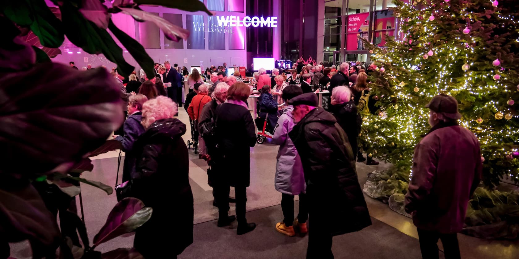 People gather in a lit hall with a large Christmas tree and a 'WELCOME' sign in the background.