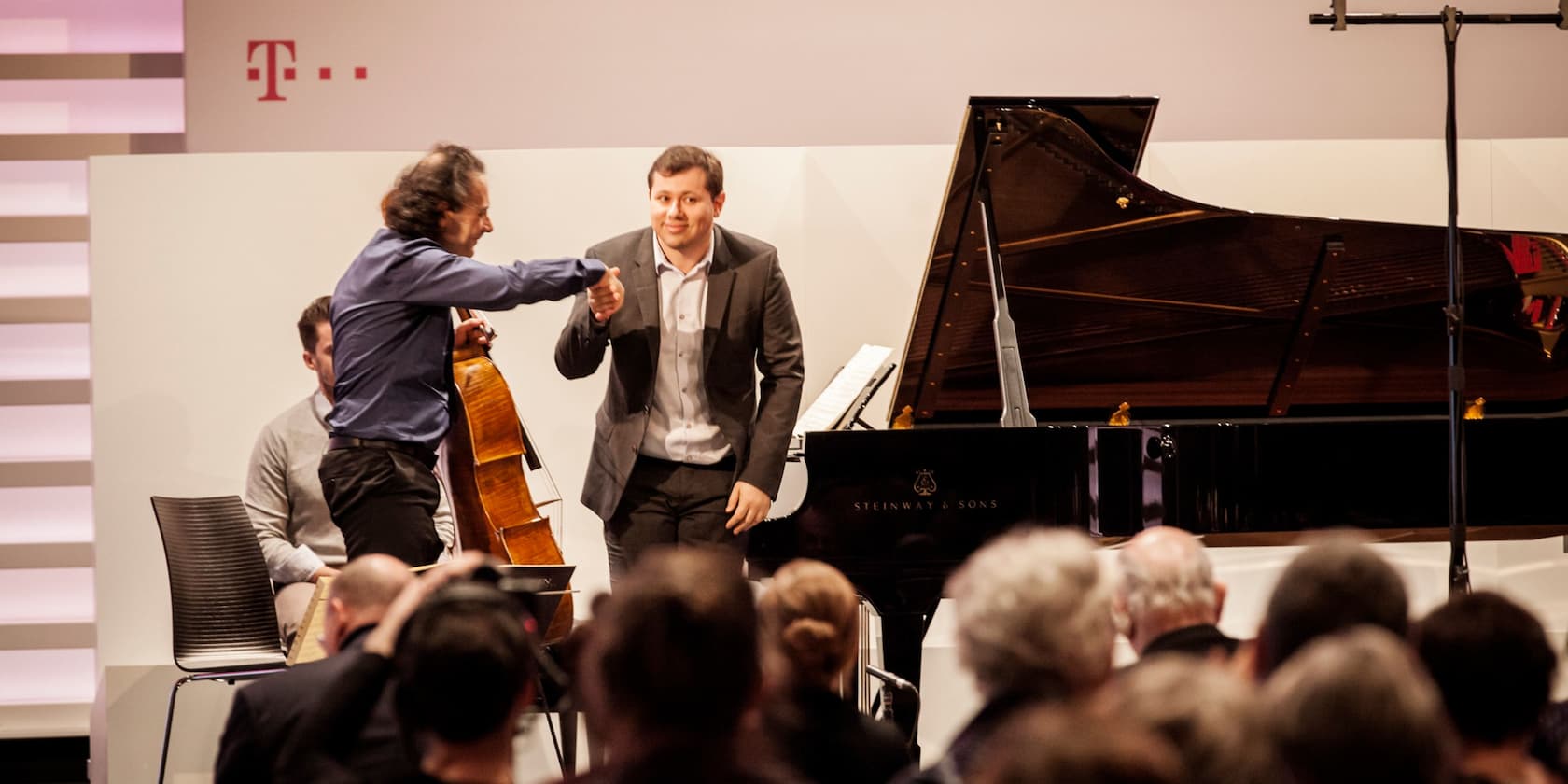 Two men bow and shake hands on stage at a concert where a grand piano and cello are present.