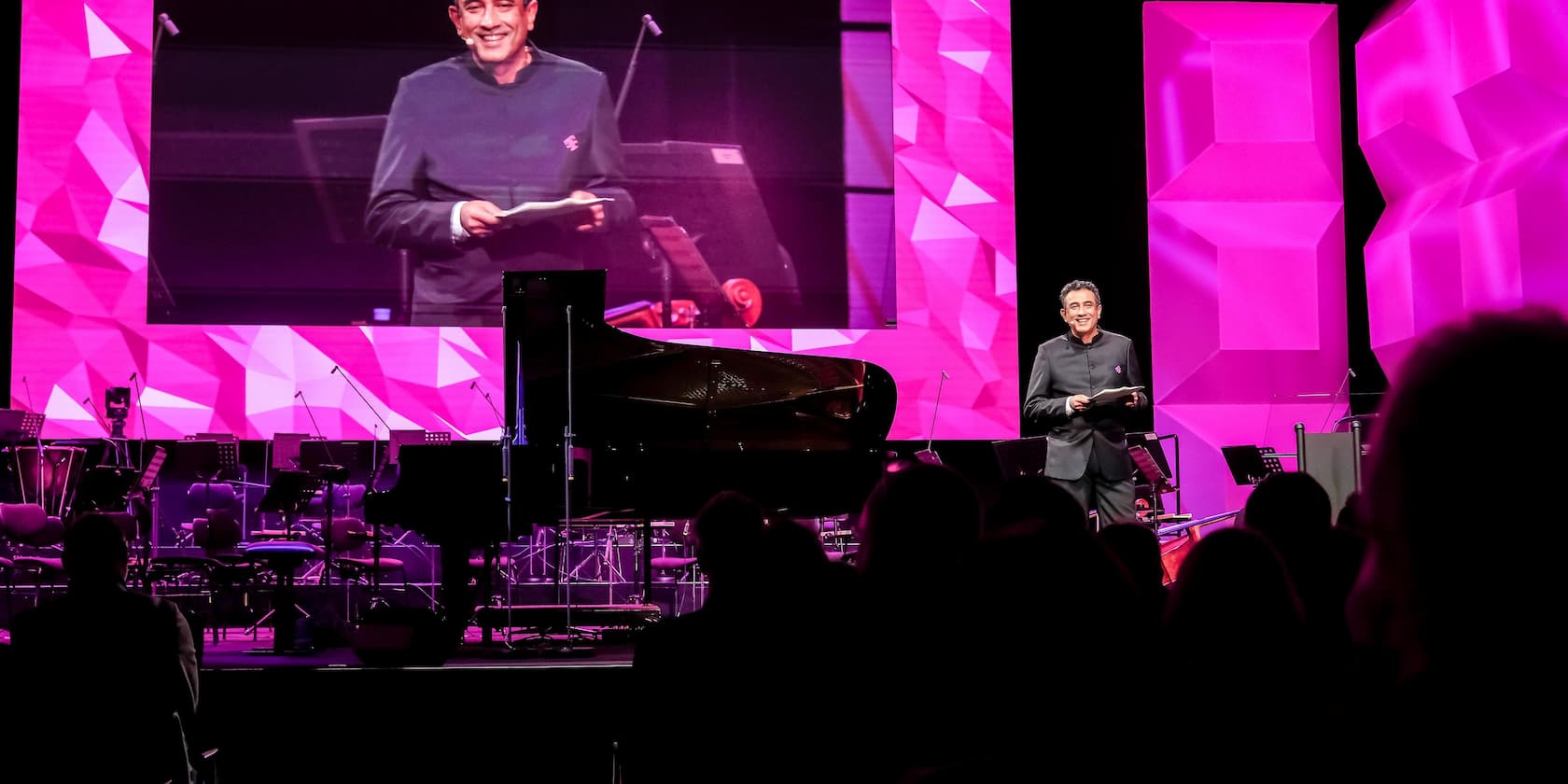 A man stands on a stage with a piano and speaks to an audience. Large, pink-lit screens are in the background.