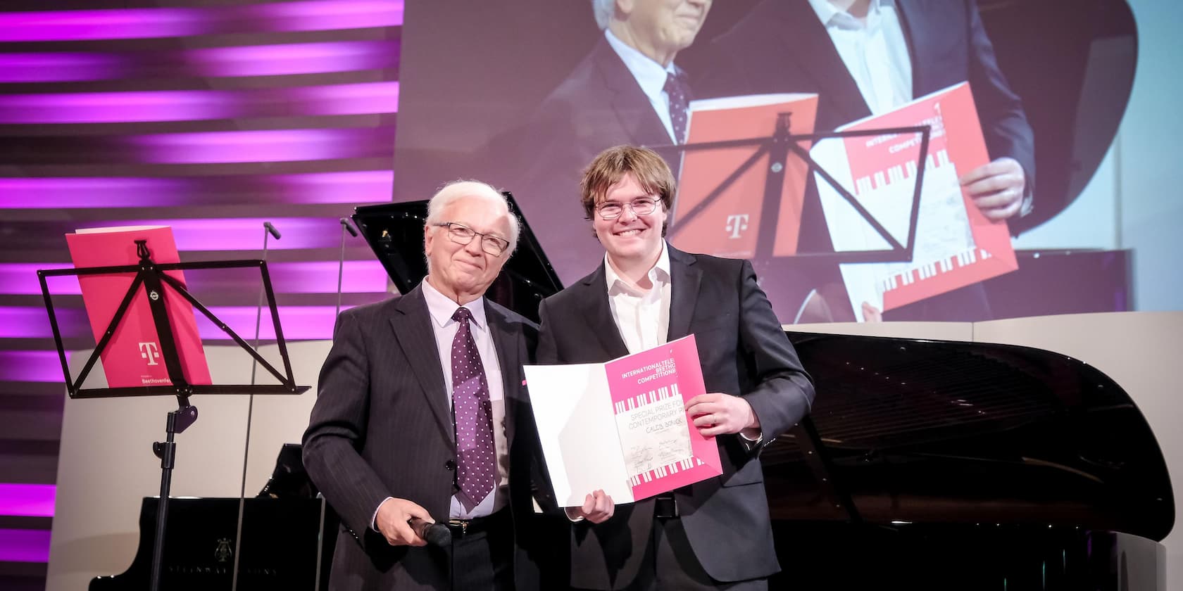 Two men stand on a stage holding a certificate. In the background, a piano and a music stand with a folder.