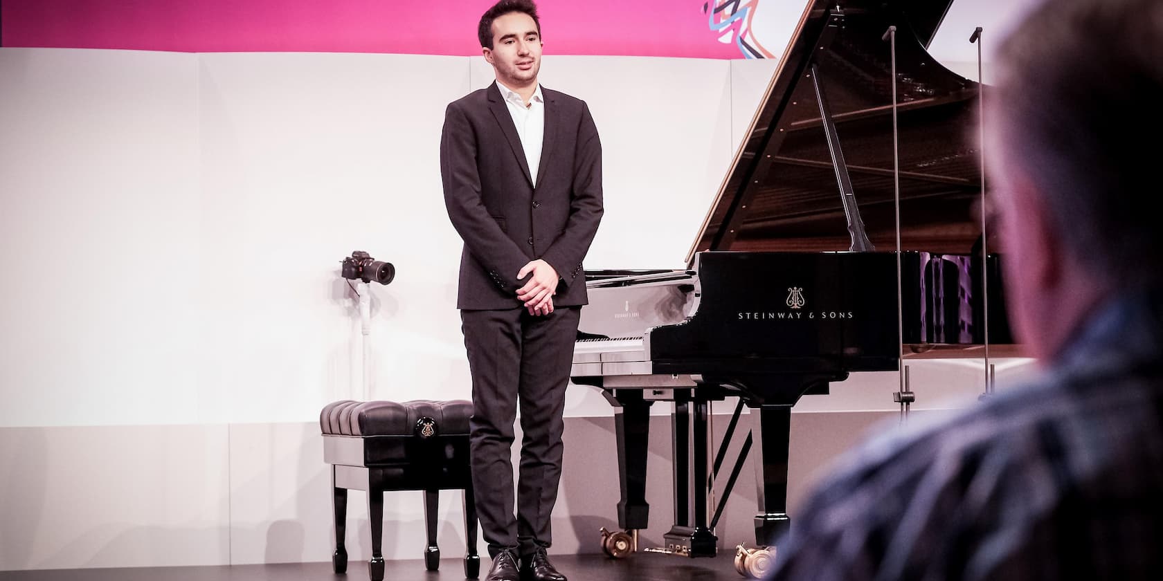 A man stands next to a Steinway & Sons grand piano on a stage, facing a small audience.