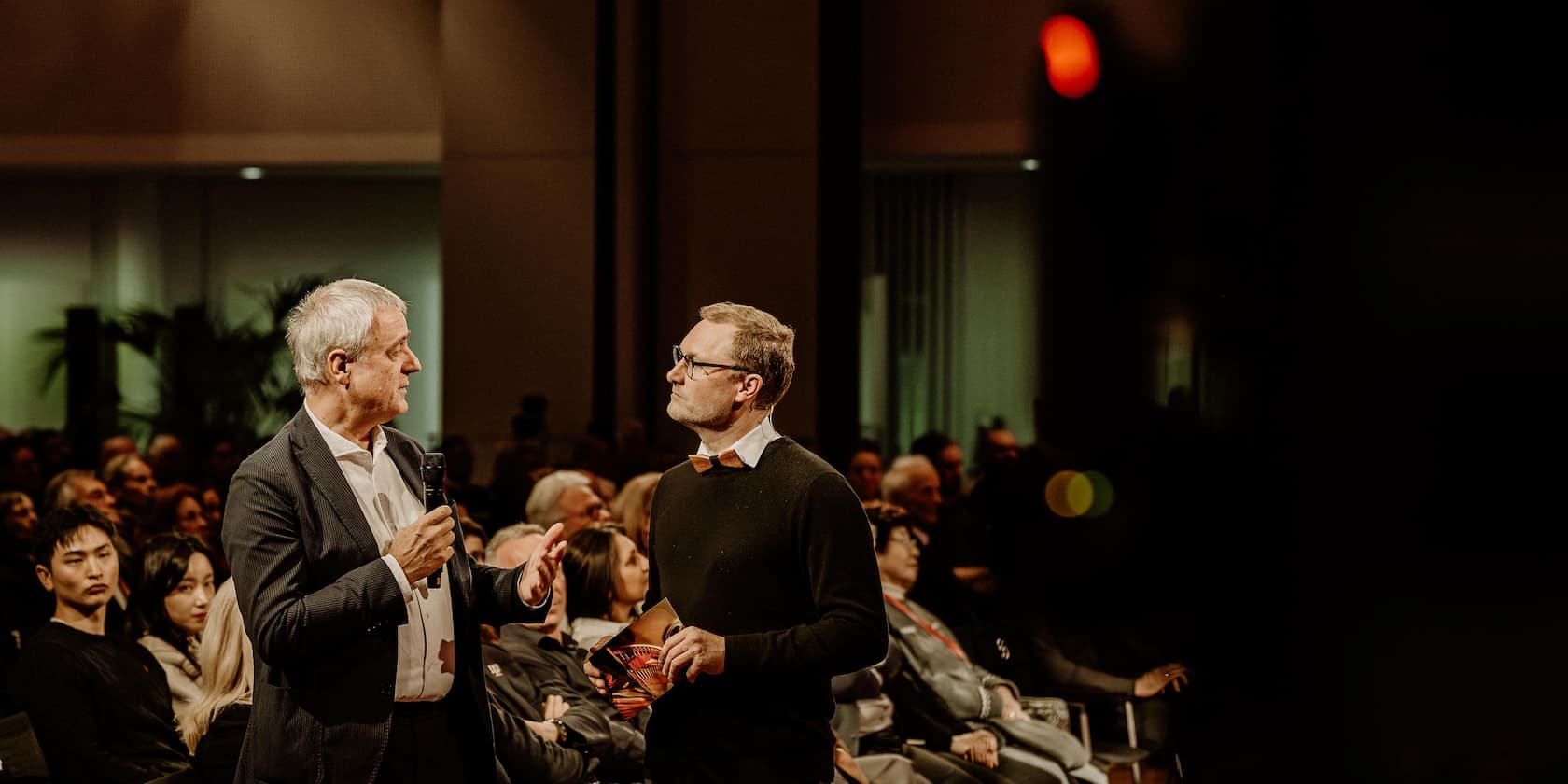 Two men discussing on a stage in front of an audience at an event.