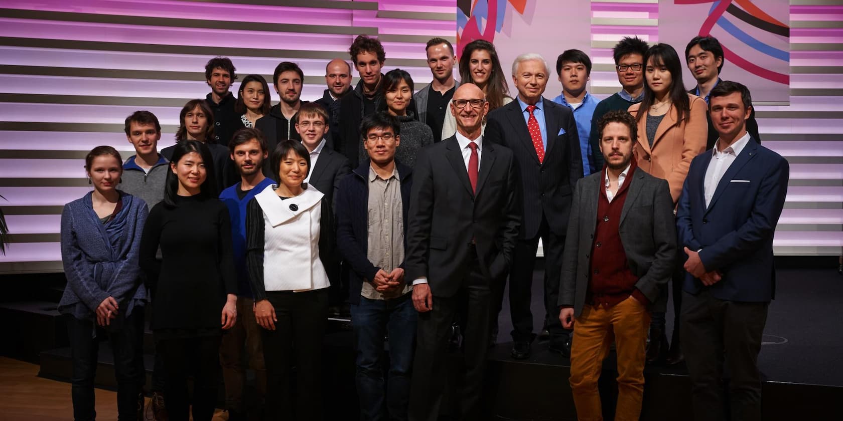 Eine Gruppe von etwa 20 Personen steht auf einer Bühne vor einem gestreiften Hintergrund mit lila Beleuchtung.