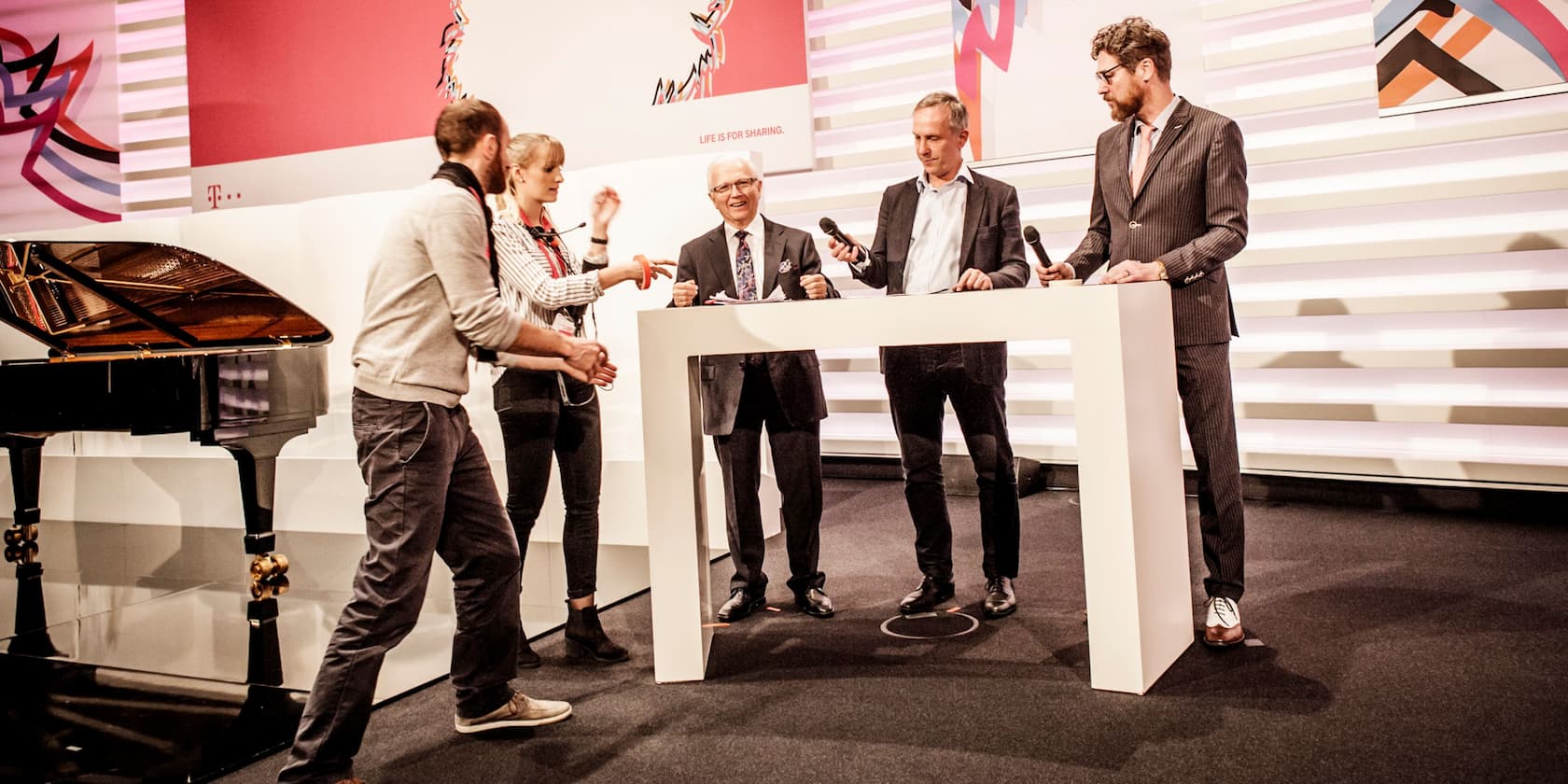 Several people stand behind a table on a stage holding microphones. In the background, there is a grand piano and marketing graphics.