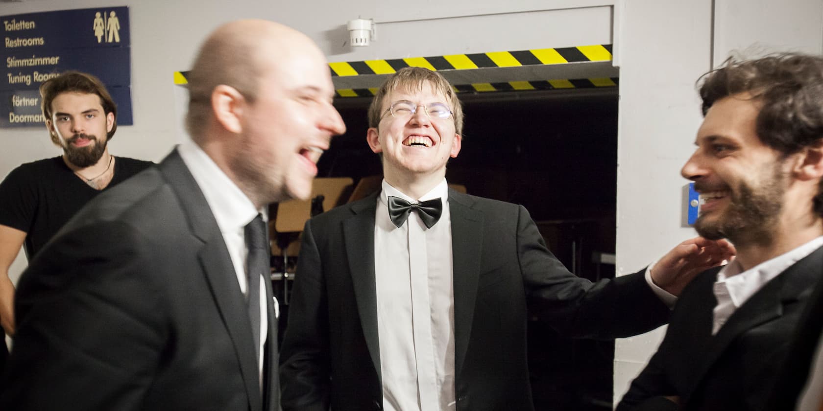 Four men in suits are standing and laughing in a hallway. A sign with restroom and other directions is visible in the background.