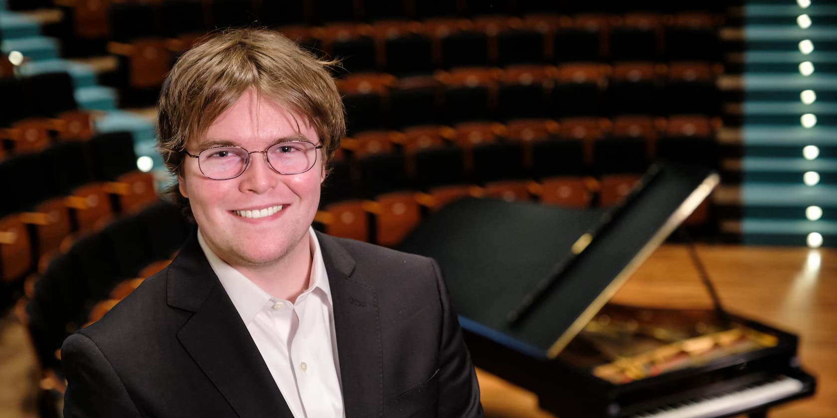 A man in a suit smiles at the camera, with a grand piano in an empty auditorium in the background.