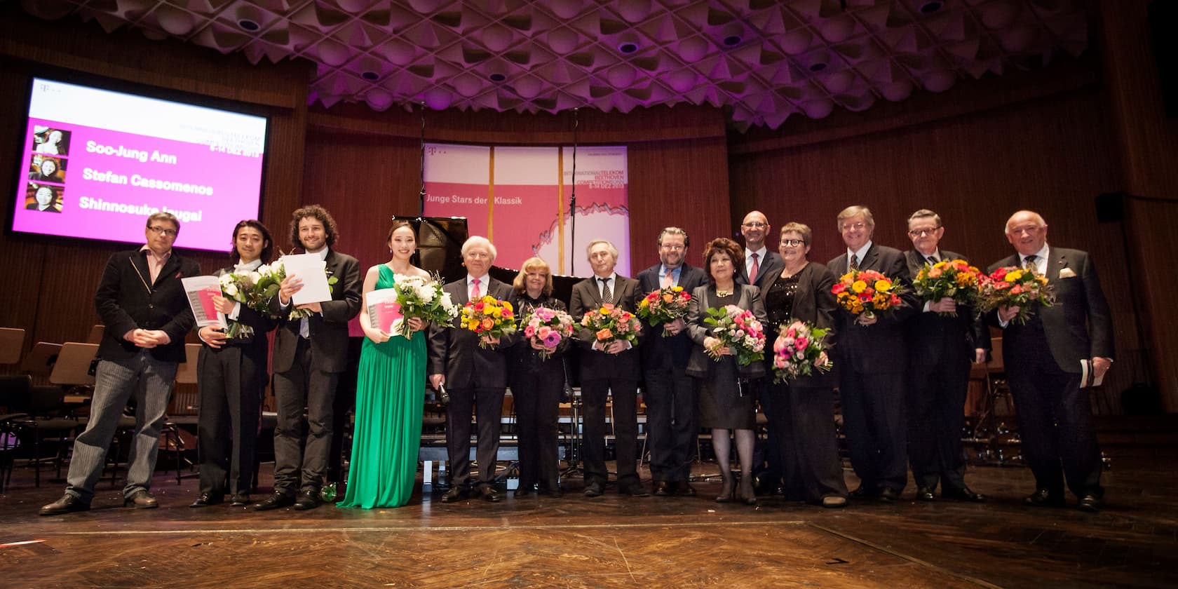 Group of people holding bouquets on stage during an award ceremony. In the background, a screen displays the names Soo-Jung Ann, Stefan Cassomenos, and Shinnosuke Inugai.