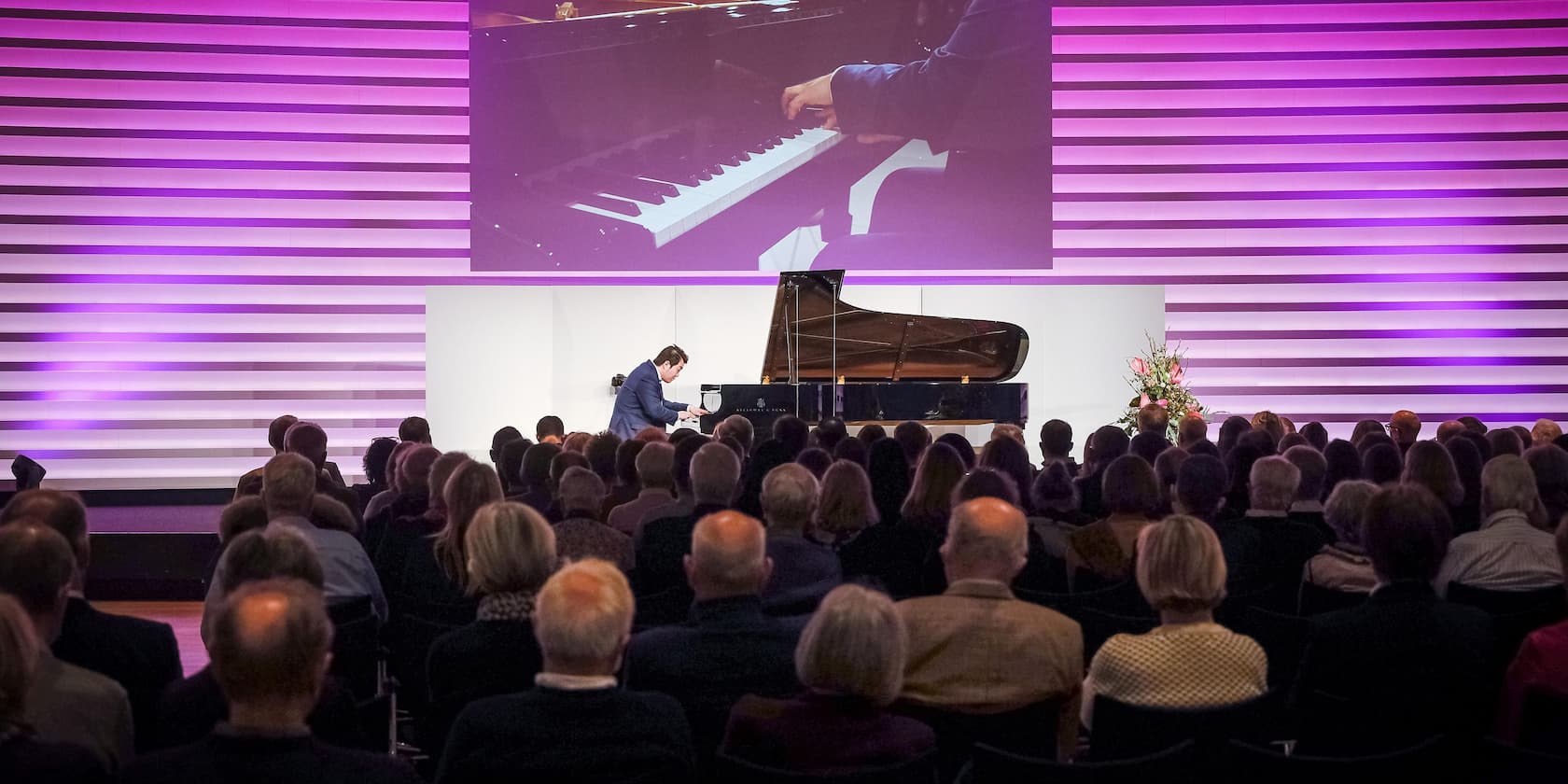 A pianist performs at a concert in front of an audience, with a projection of his hands on the keyboard in the background.