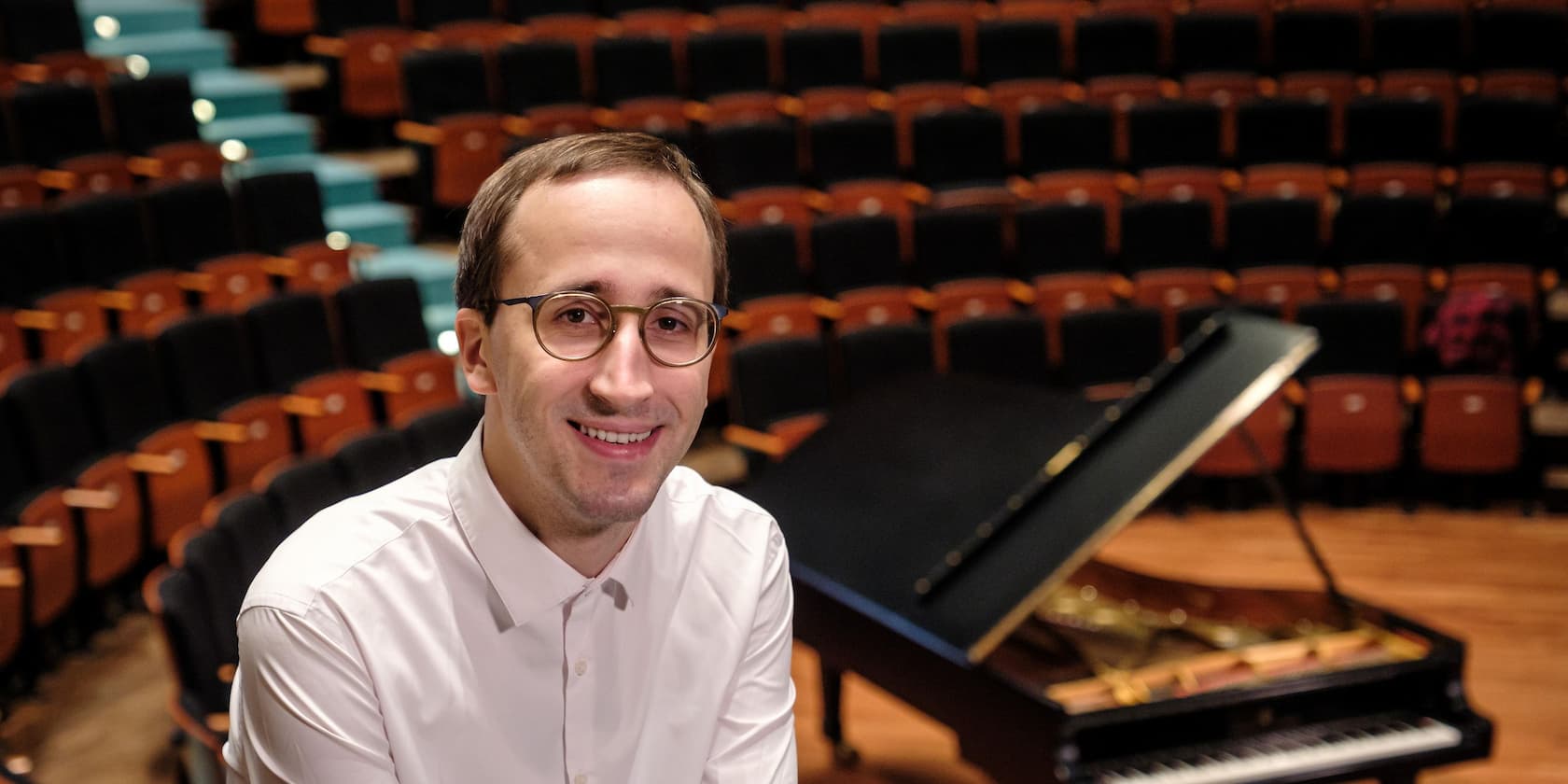 Person with glasses and a white shirt sitting in front of a grand piano in an empty concert hall.