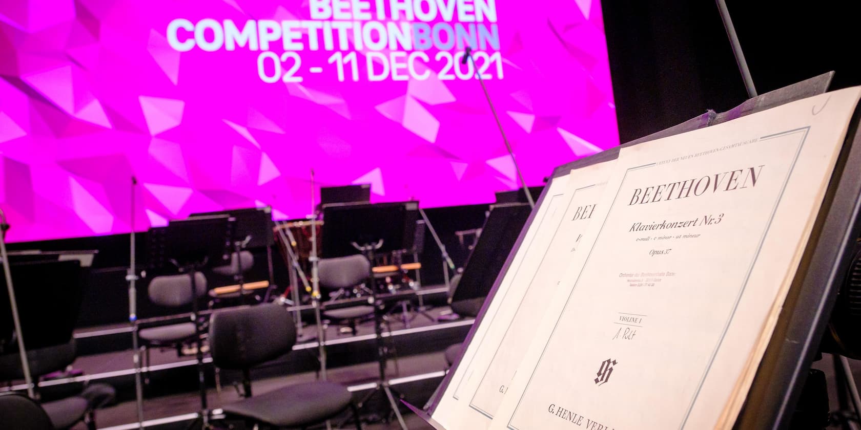 Notenständer mit Partitur von Beethovens Klavierkonzert Nr. 3, im Hintergrund leere Stühle und 'Beethoven Competition Bonn 02 - 11 Dec 2021' auf rosa Bildschirm.