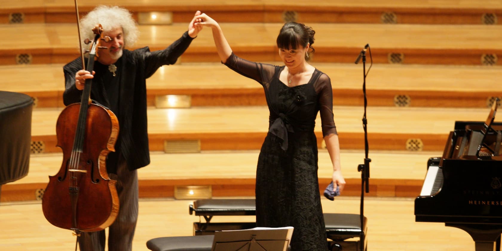 Two musicians performing on stage, a cellist and a pianist holding hands.