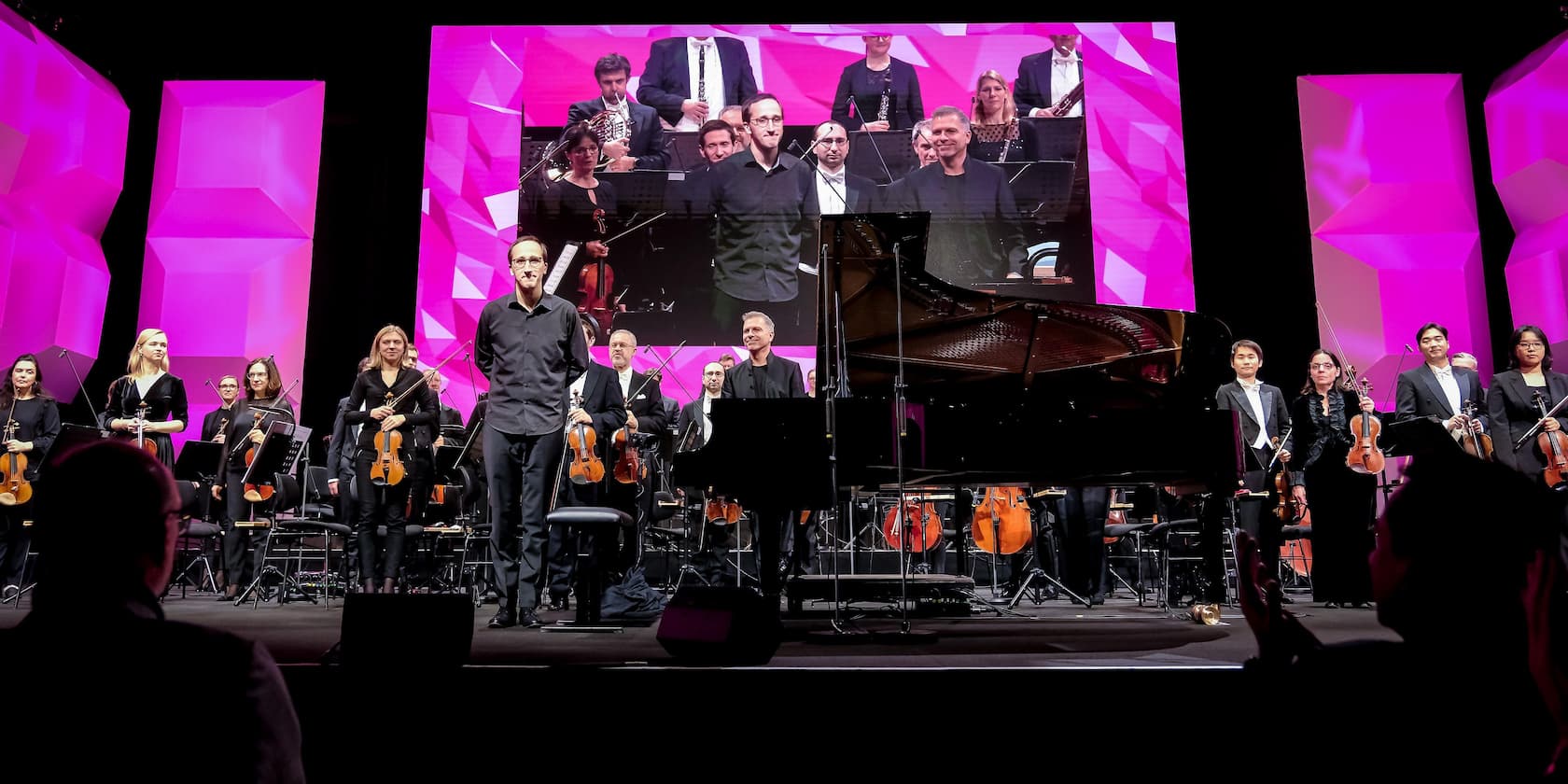 An orchestra stands on a stage with a large screen in the background. The screen shows a close-up of the musicians. The stage is surrounded by pink geometric structures.