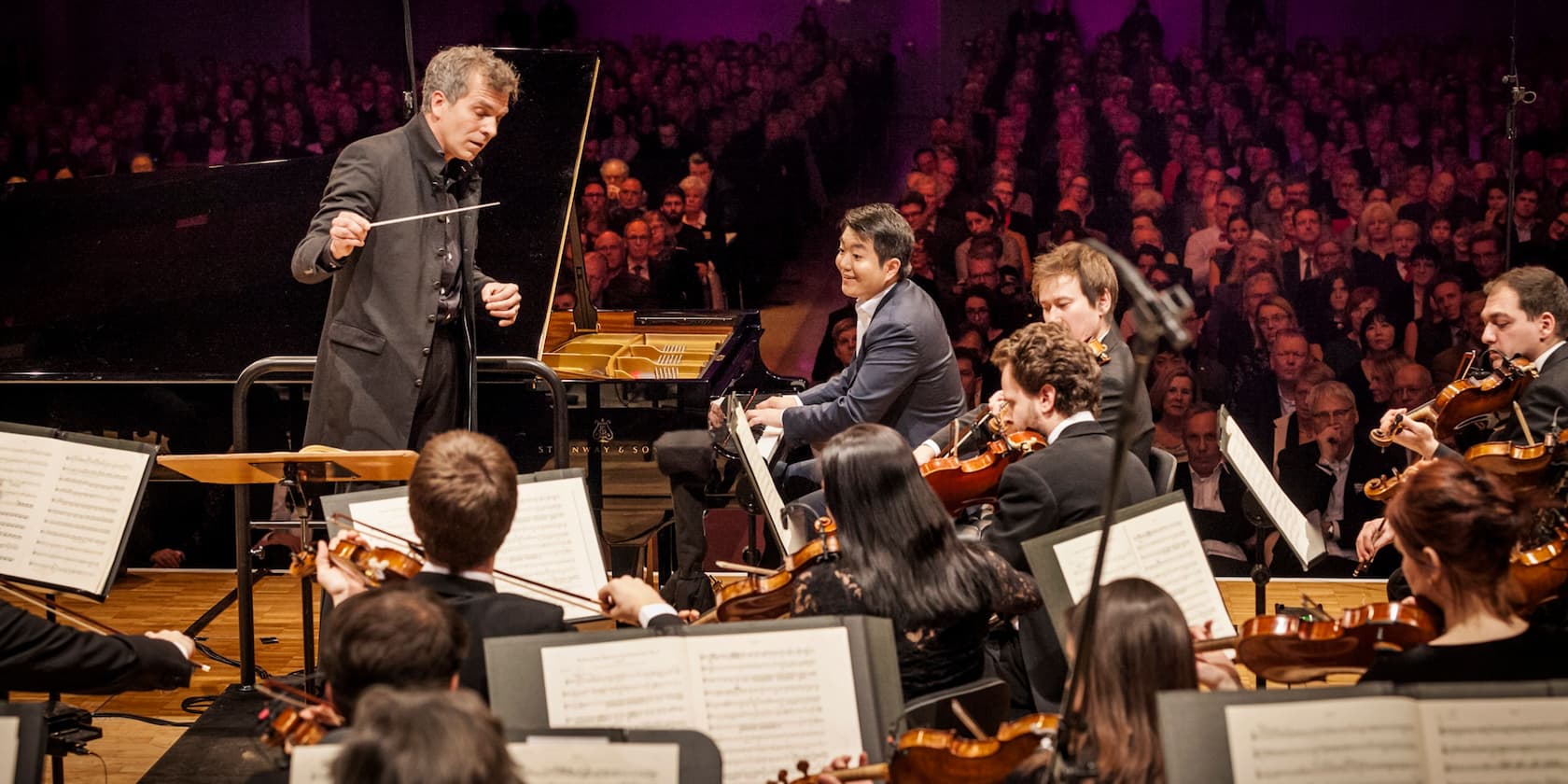 Conductor leading an orchestra while a pianist plays in the background. Audience in the concert hall.