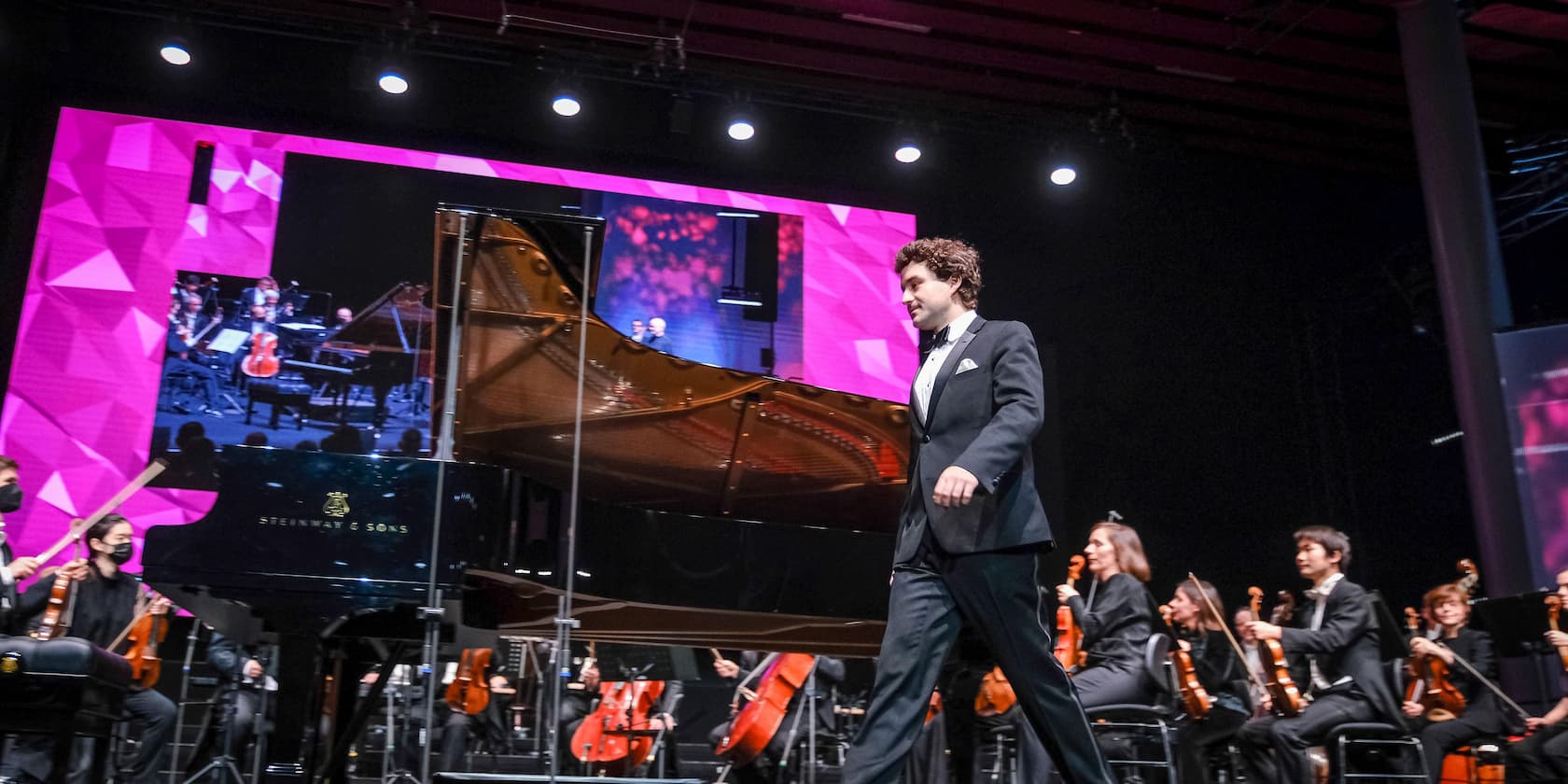 A concert pianist exits the stage after a performance with an orchestra. A large screen displaying a live feed is visible in the background.