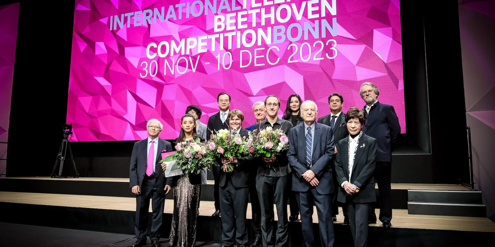 Group posing on stage in front of a sign for the International Beethoven Competition Bonn 2023 with bouquets of flowers.