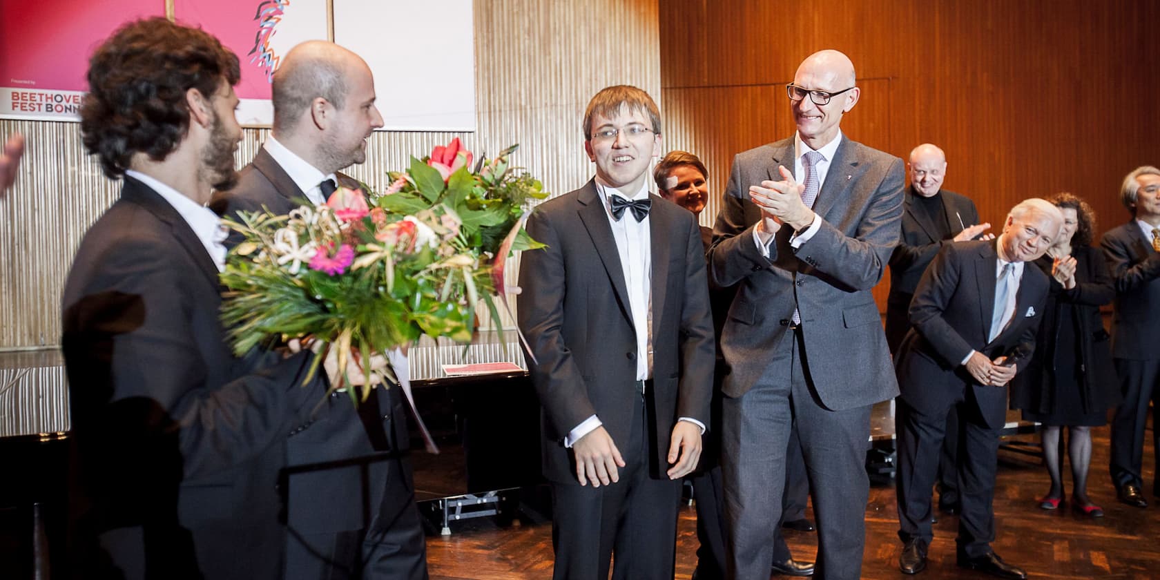 A young man in a suit receives a bouquet of flowers at an event while several people in formal attire applaud him.