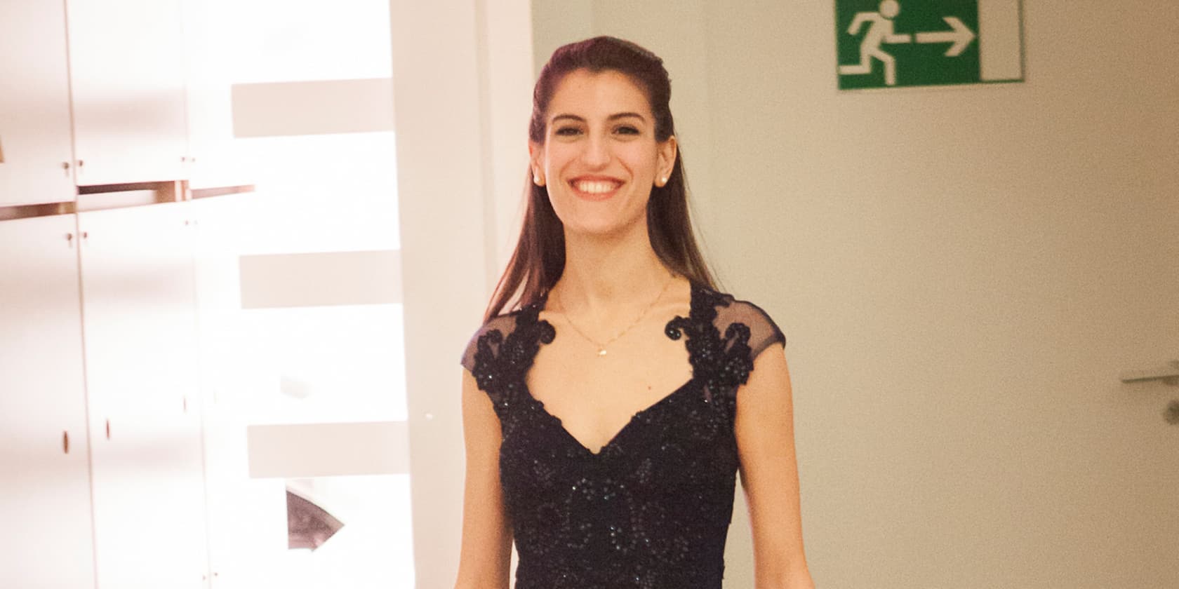 Woman in an elegant black dress smiling, standing next to an emergency exit sign.