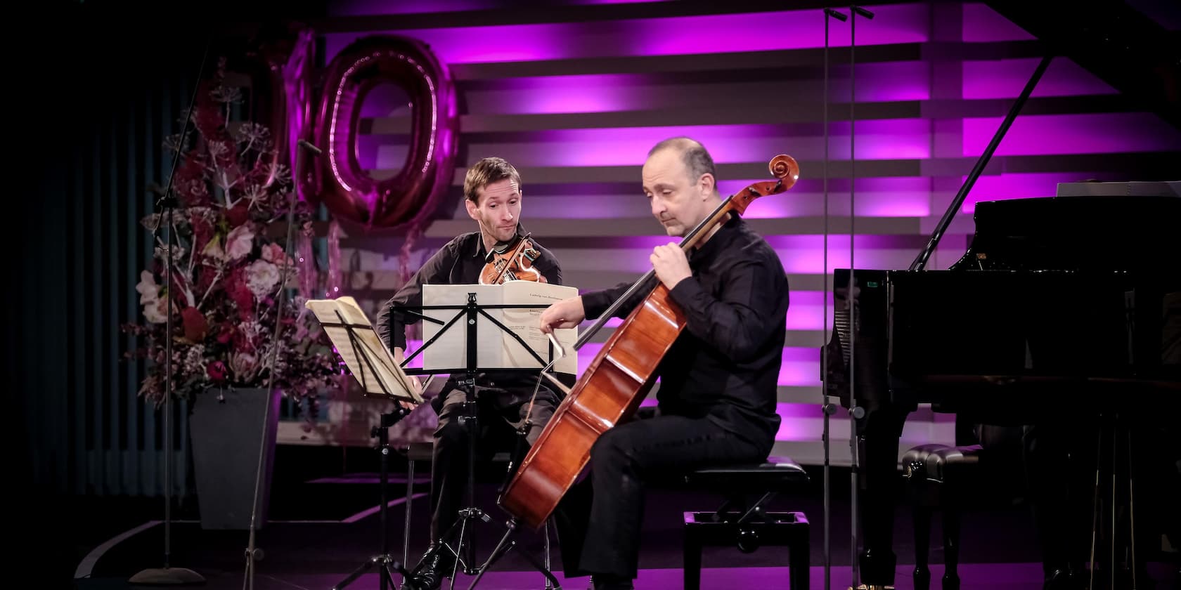 Two musicians playing violin and cello on a stage with purple background lighting. In the background, there are flower arrangements and the number 10 in balloons.