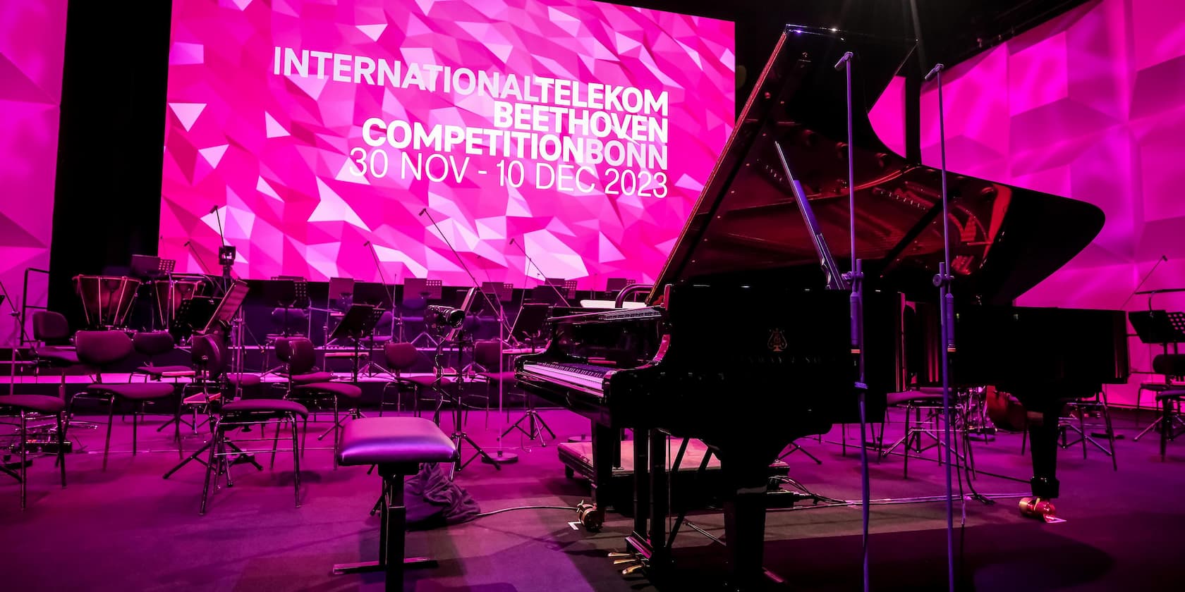 Konzertbühne mit einem Flügel und mehreren Stühlen vor einem pinkfarbenen Hintergrund mit der Aufschrift 'INTERNATIONAL TELEKOM BEETHOVEN COMPETITION BONN 30 NOV - 10 DEC 2023'.