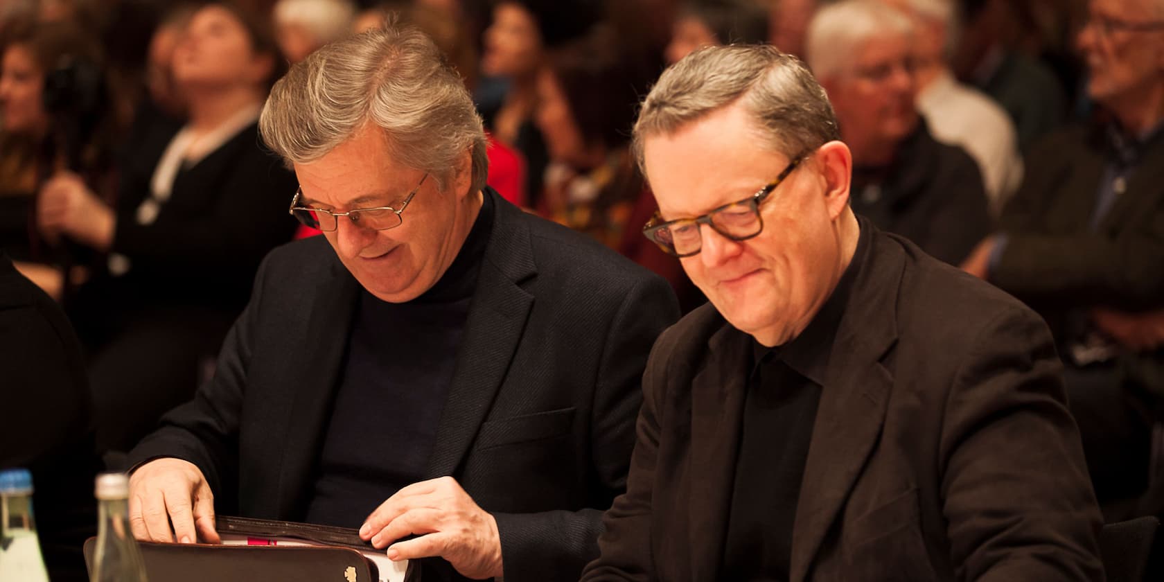 Two older men sitting at a table reading during an event.