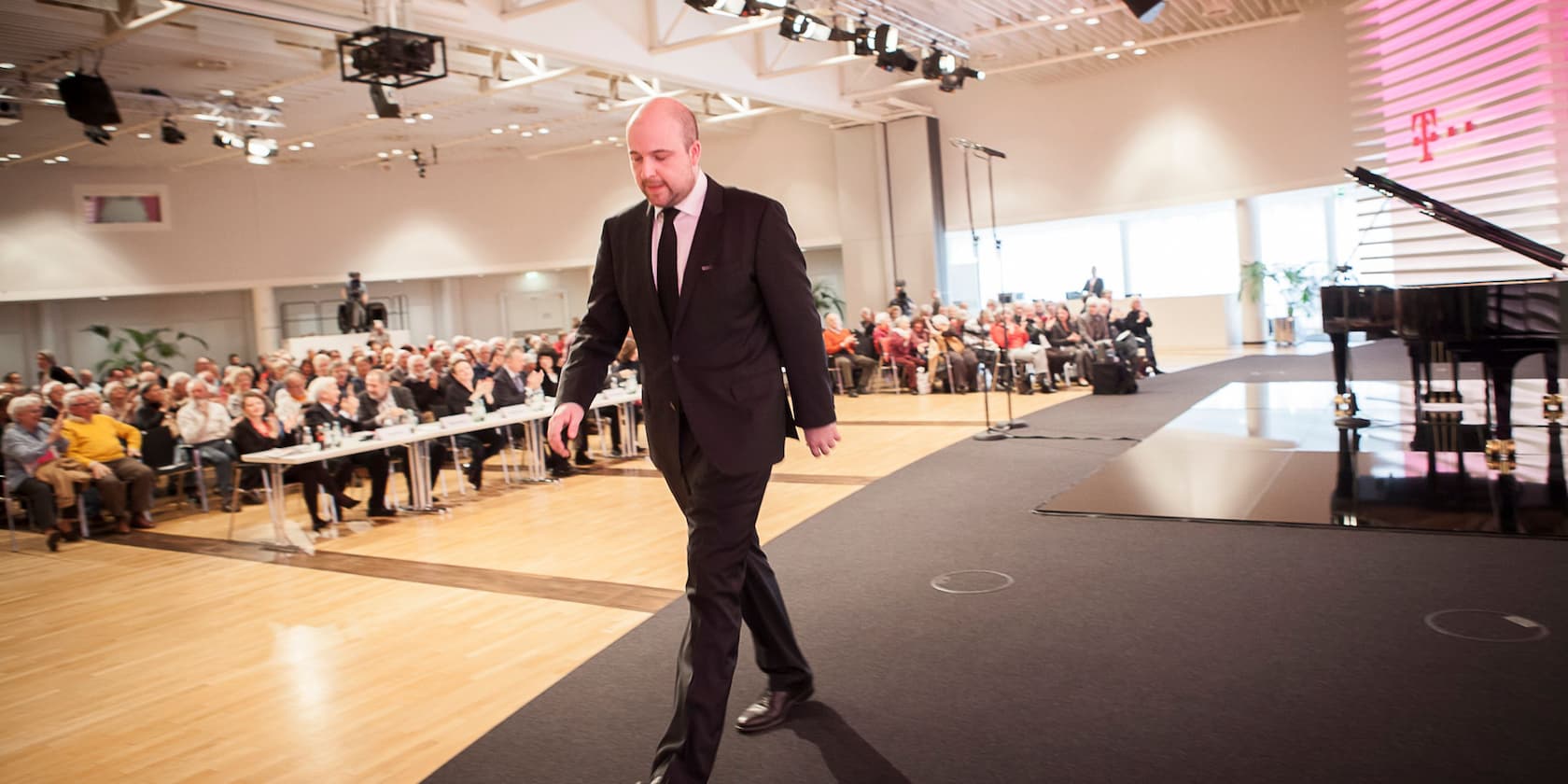 A man in a suit walking on a stage, while an audience sits at tables and listens.