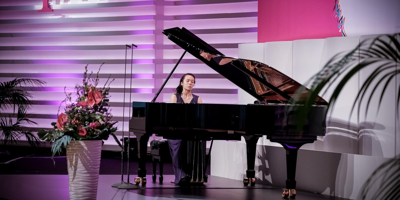 A woman plays the piano on a stage. A vase with flowers is in the foreground. The stage is lit in purple and white.