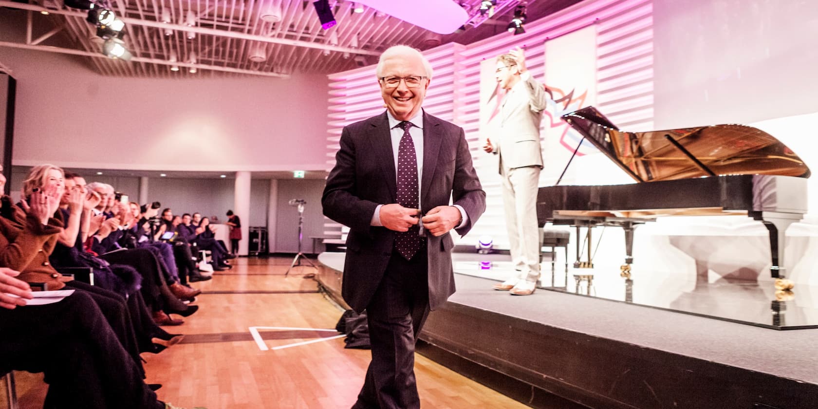 A man in a suit on a stage smiles as the audience applauds. A grand piano is visible in the background.