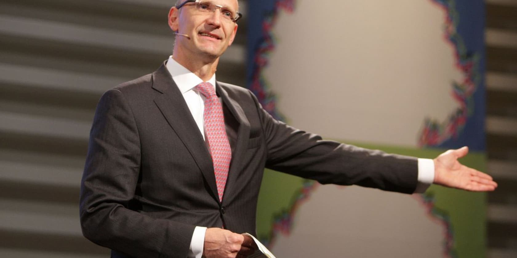 A man in a suit and tie speaking on a stage with an outstretched arm.