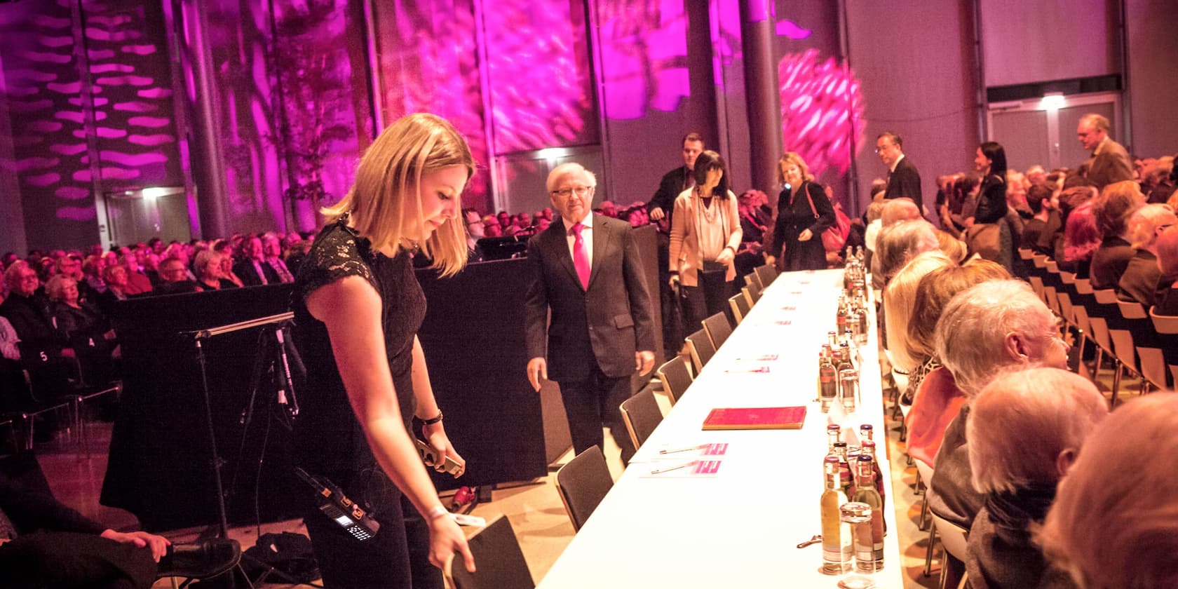 Several people are at an event in a room with pink lighting. A woman in the foreground is arranging something, while others are seated or standing in the background.