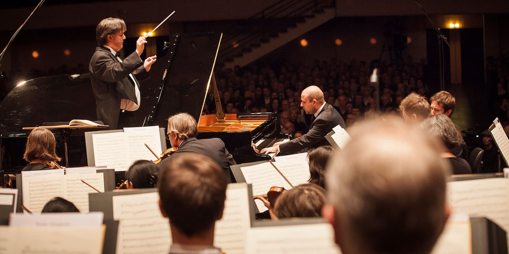 A conductor leads an orchestra while a pianist performs, in front of an audience in a concert hall.