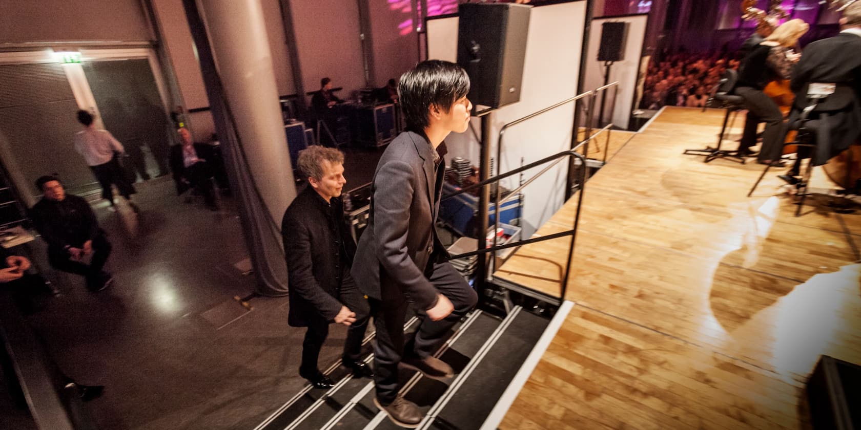 Two men walk up stairs towards a stage while an orchestra performance takes place in the background.