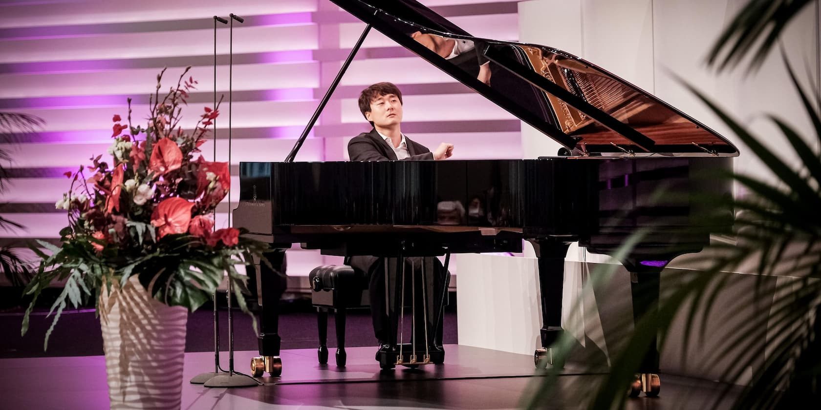 A man playing the piano on a stage, with a flower arrangement in the foreground.