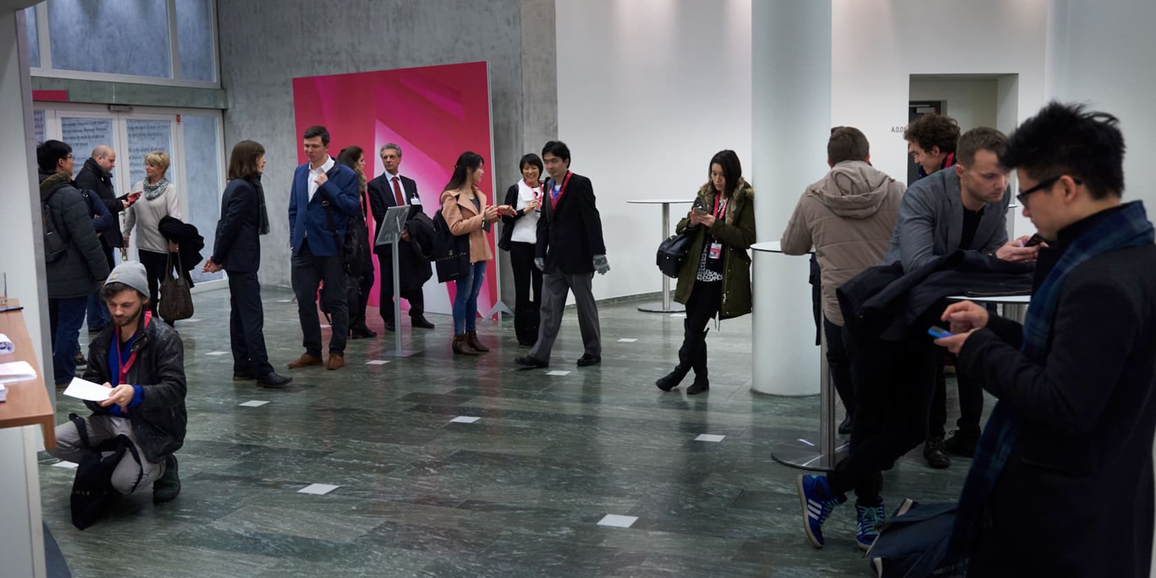 People gather in the lobby of a modern building, some conversing while others look at their smartphones.