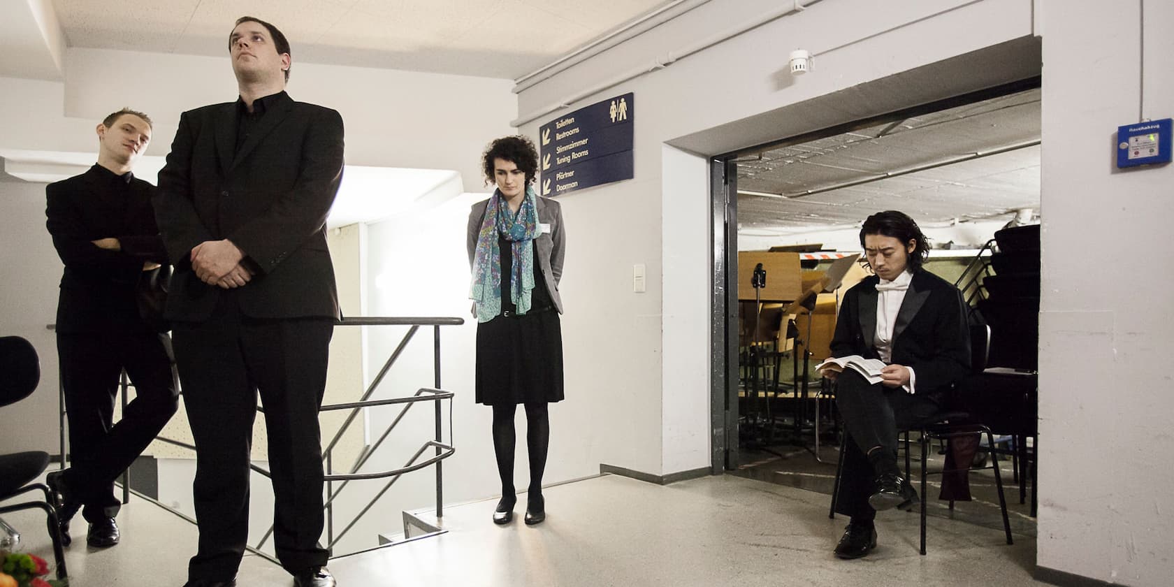 Four people in formal clothing are waiting indoors. One is sitting on a chair reading a book.