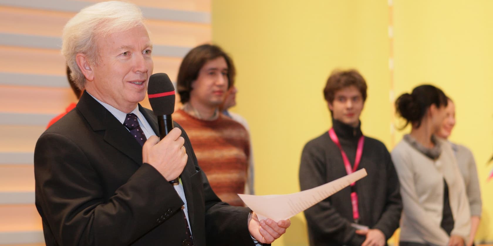 An elderly man in a suit speaks into a microphone while holding a paper. Several people stand in the background.