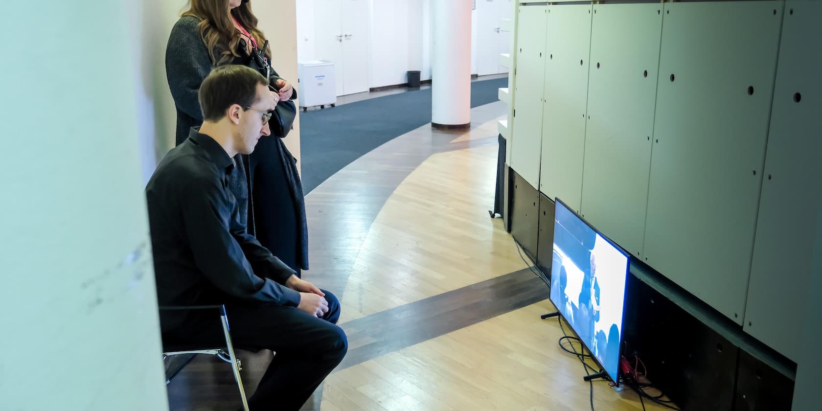 Two people standing and one person sitting on a chair while watching a presentation on a screen placed on the floor.