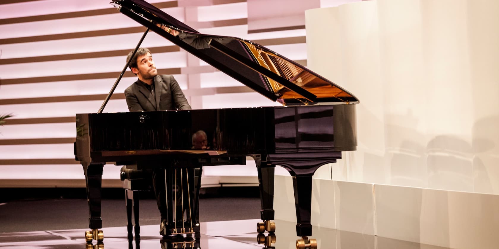 A man plays on a black grand piano in front of a striped background.