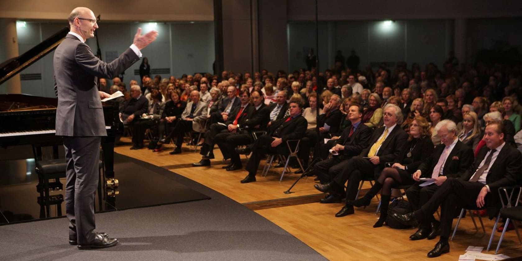 A man gives a speech to an audience in a hall. A grand piano is in the background.