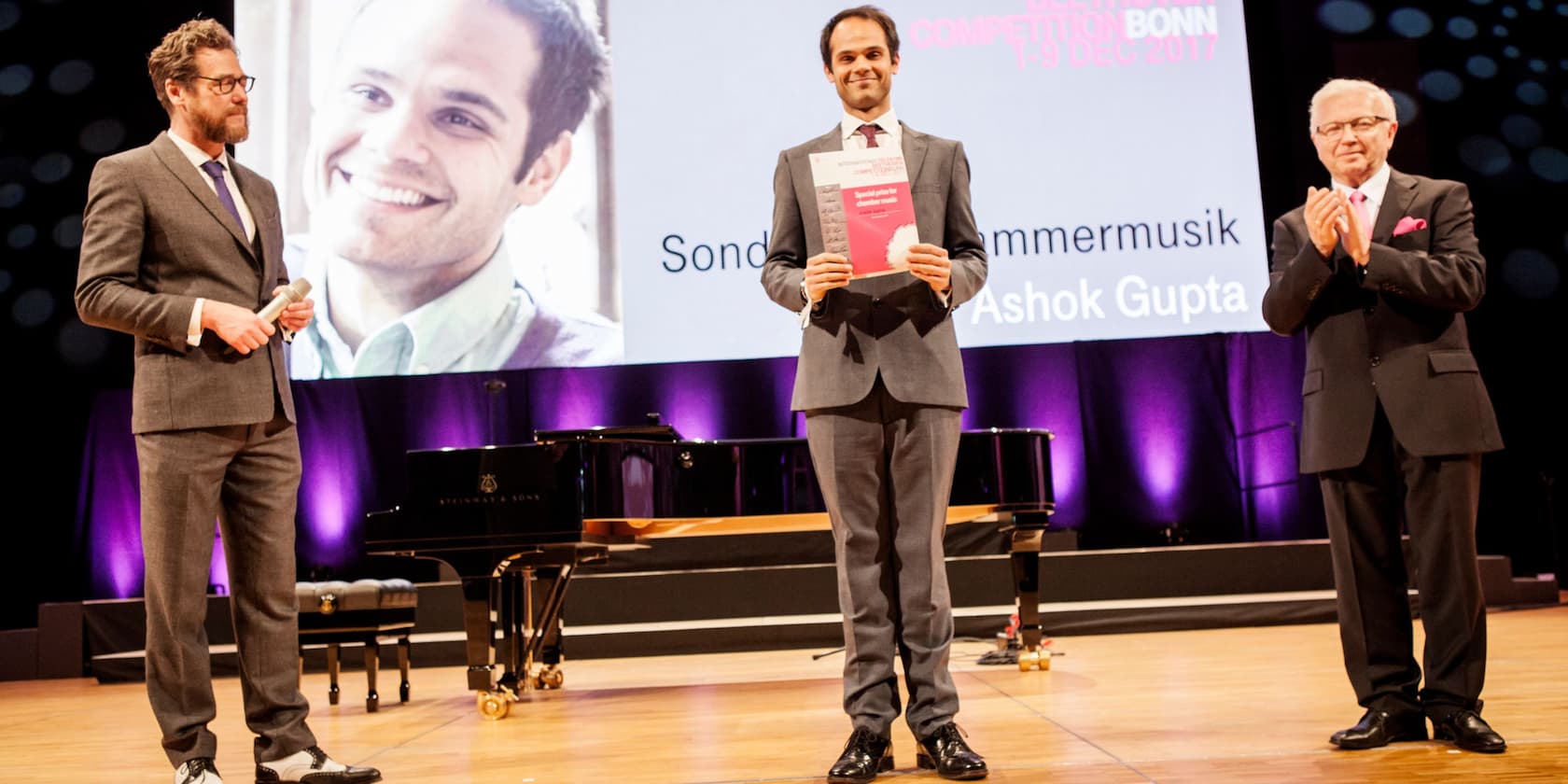 Concert hall with three men in suits on stage. One of the men holds a certificate reading 'Special prize for chamber music' with the name Ashok Gupta. In the background, a large projected image and a grand piano.