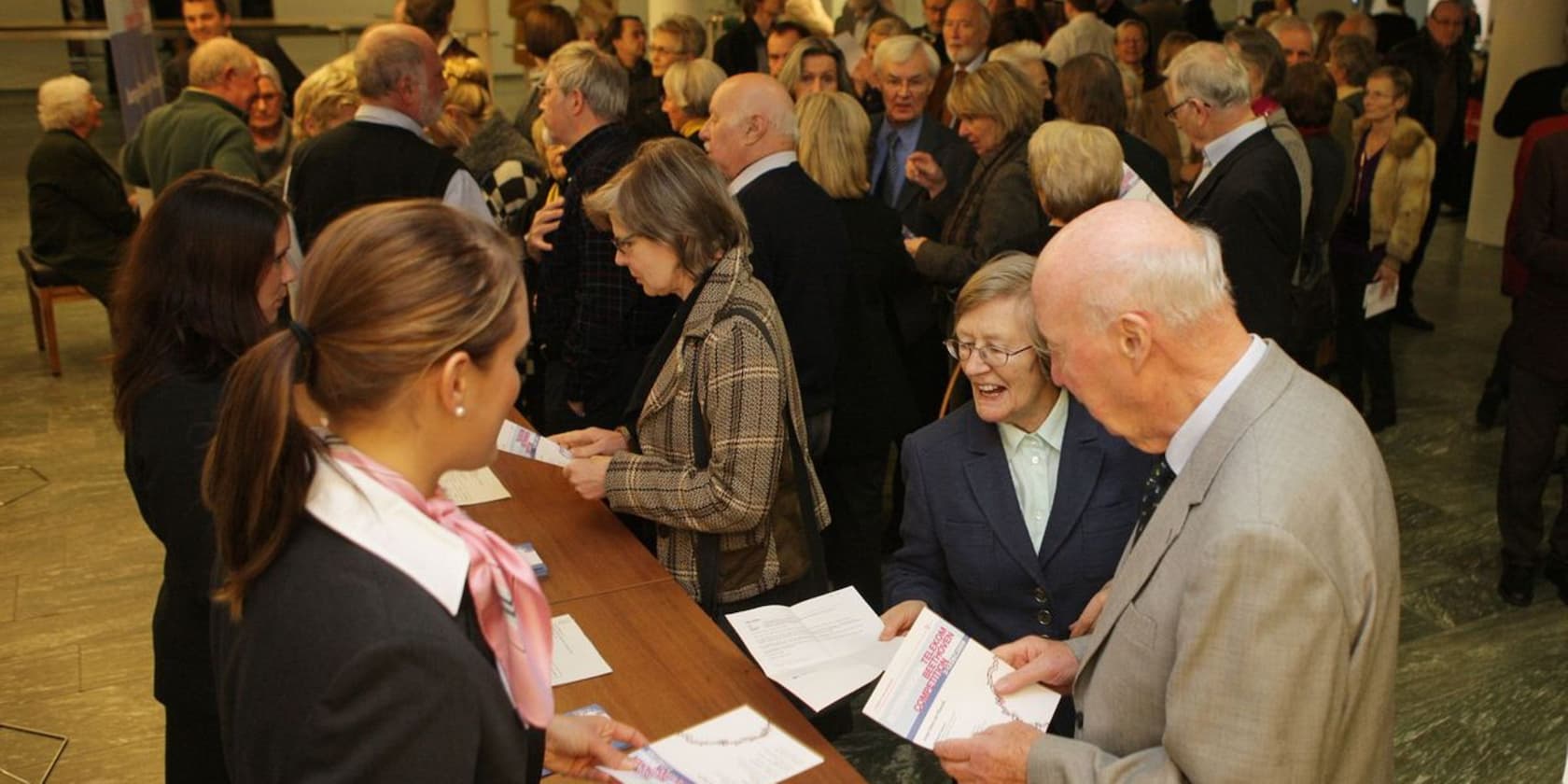 A crowd of people at an event, some talking to each other and holding papers.