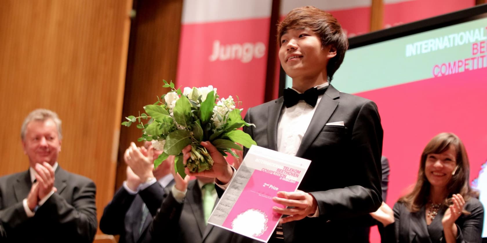 Young man in a suit holding a certificate and a bouquet of flowers, surrounded by applauding people on stage.