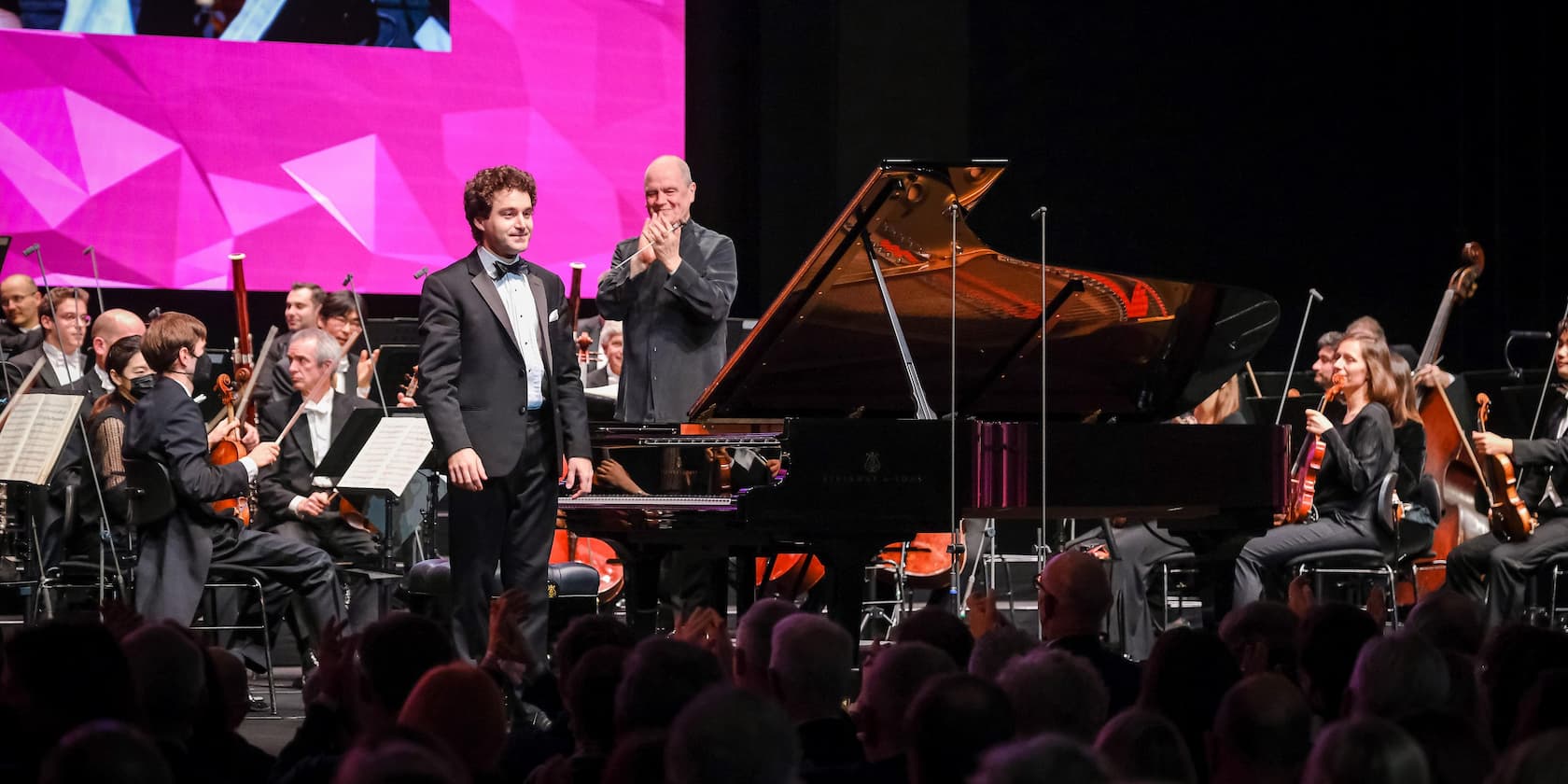 A pianist takes a bow in front of the audience while an orchestra applauds in the background.