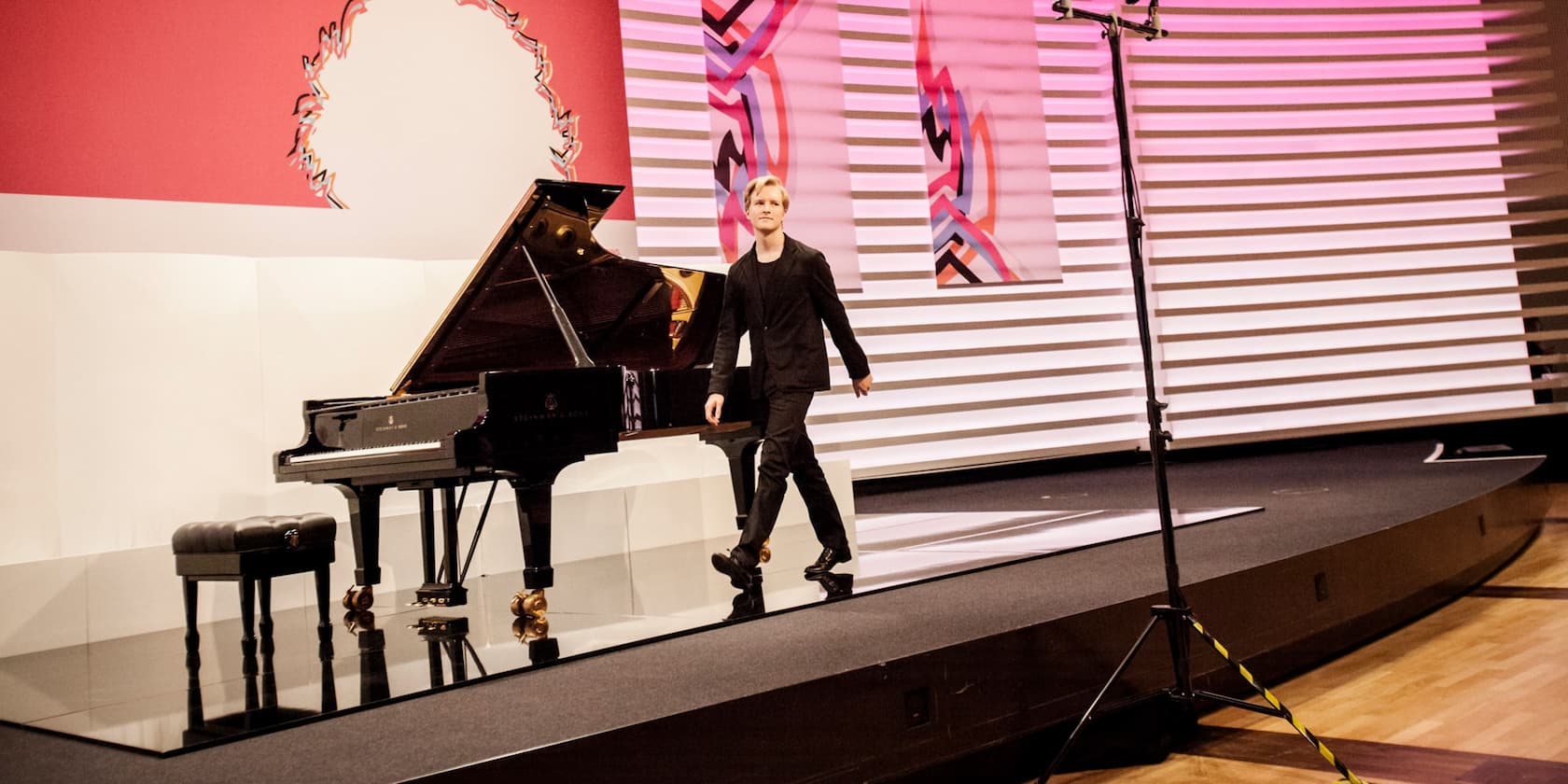 A man in black clothing walks along a stage. A black grand piano is placed on the stage.