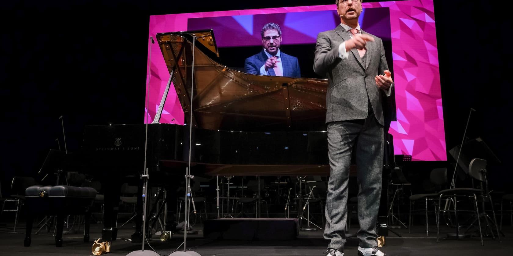 A man in a suit stands on a stage in front of a piano, with a large screen showing his image in the background.