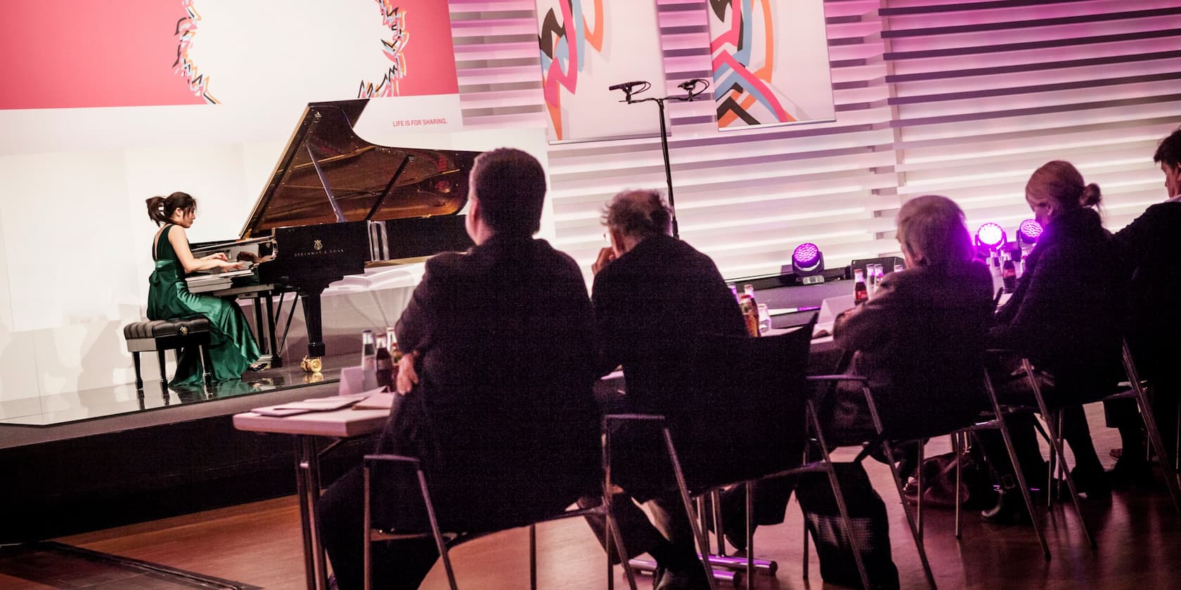 A woman in a green dress plays the piano on a stage in front of a seated audience.