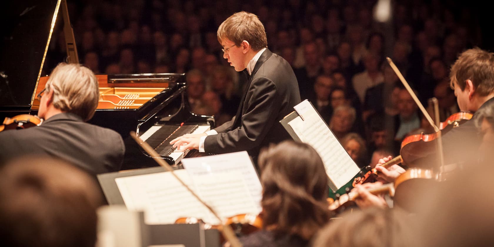 Konzertpianist spielt am Klavier, umgeben von einem Orchester auf der Bühne.